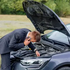 Mężczyzna w garniturze rozmawia przez telefon, pochylając się nad otwartą maską samochodu.