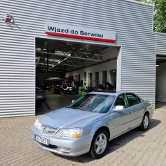 Srebrny sedan przed wjazdem do serwisu. W tle widać motocykle i sprzęt warsztatowy. Może to miejsce, gdzie naprawią mój carprog2?