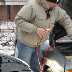 Mężczyzna w zimowej kurtce podłącza kable rozruchowe do silnika samochodu w zimowy dzień.