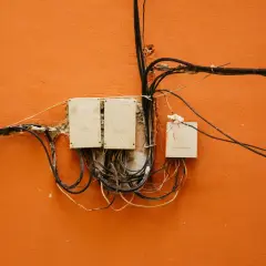 Stare skrzynki elektryczne z plątaniną kabli na pomarańczowej ścianie.