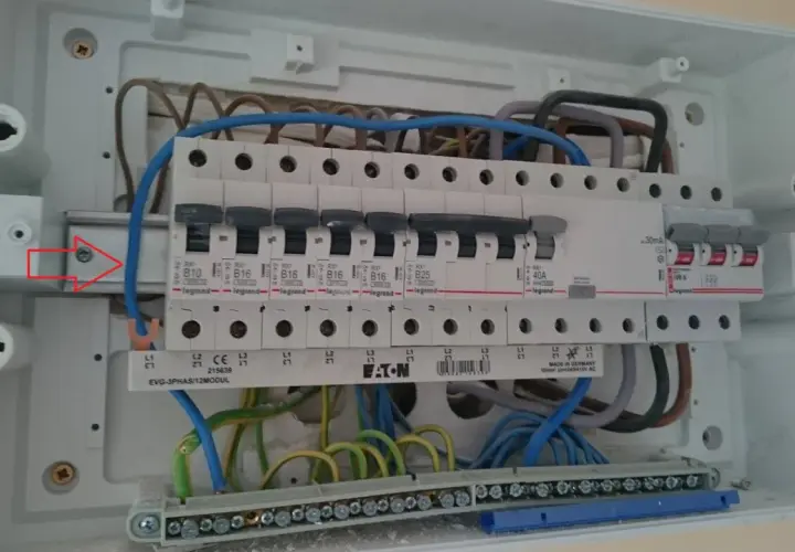 W skrzynce elektrycznej widać zabezpieczenie przedlicznikowe, bezpieczniki i przewody. Czerwona strzałka wskazuje śrubę mocującą szynę.