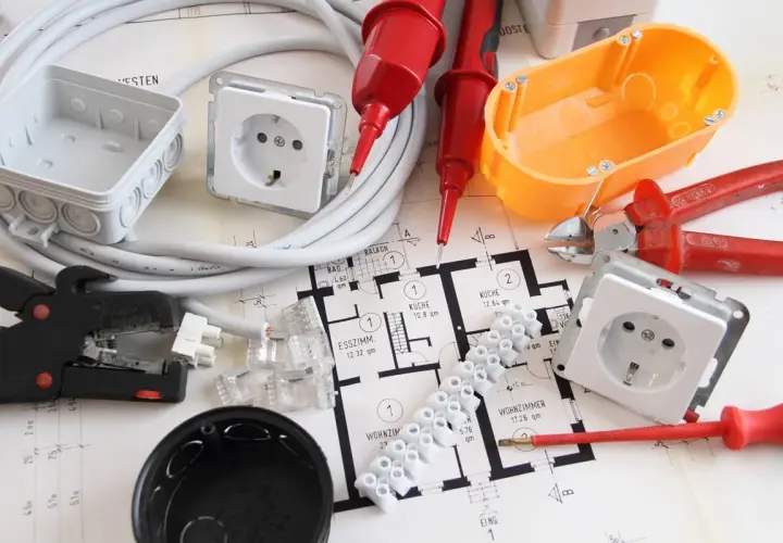 Narzędzia i materiały do wymiany instalacji elektrycznej bez kucia: przewody, puszki, gniazdka, zaciski, kombinerki, śrubokręt, miernik napięcia i plan mieszkania.