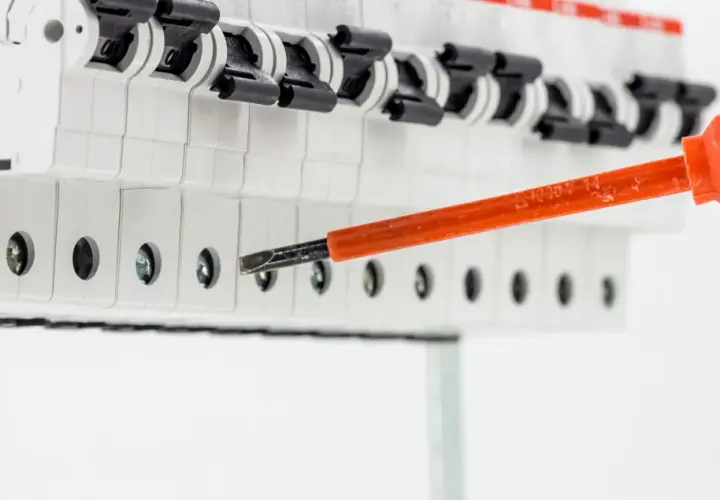 Elektryk dokręca śrubę w skrzynce z bezpiecznikami, przygotowując instalację. Widać przewód uziemienia i narzędzie rcd.