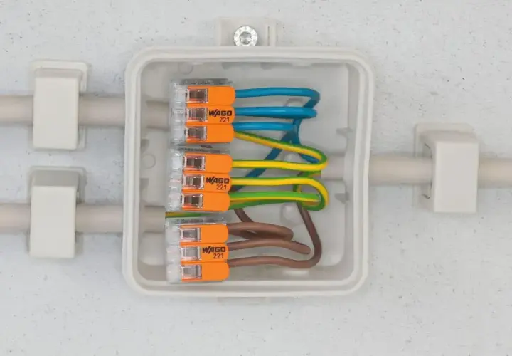 Bezpieczne przyłącze elektryczne z przewodami w kolorach niebieskim, żółto-zielonym i brązowym, połączonymi złączkami WAGO 221 w białej puszce.