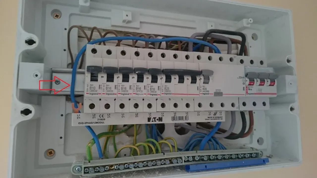 W skrzynce elektrycznej widać zabezpieczenie przedlicznikowe, bezpieczniki i przewody. Czerwona strzałka wskazuje śrubę mocującą szynę.