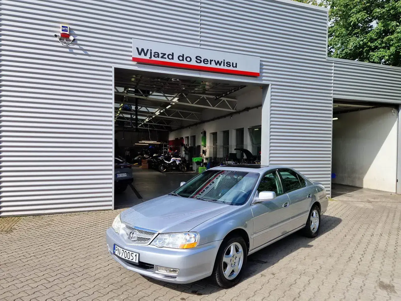 Srebrny sedan przed wjazdem do serwisu. W tle widać motocykle i sprzęt warsztatowy. Może to miejsce, gdzie naprawią mój carprog2?