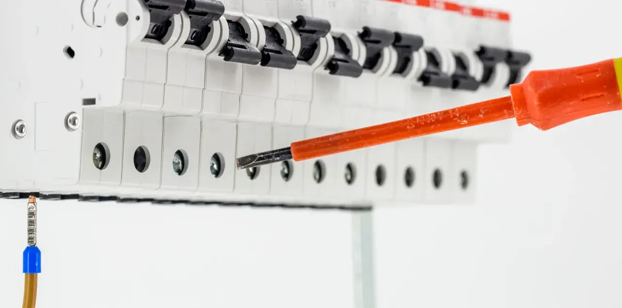 Elektryk dokręca śrubę w skrzynce z bezpiecznikami, przygotowując instalację. Widać przewód uziemienia i narzędzie rcd.