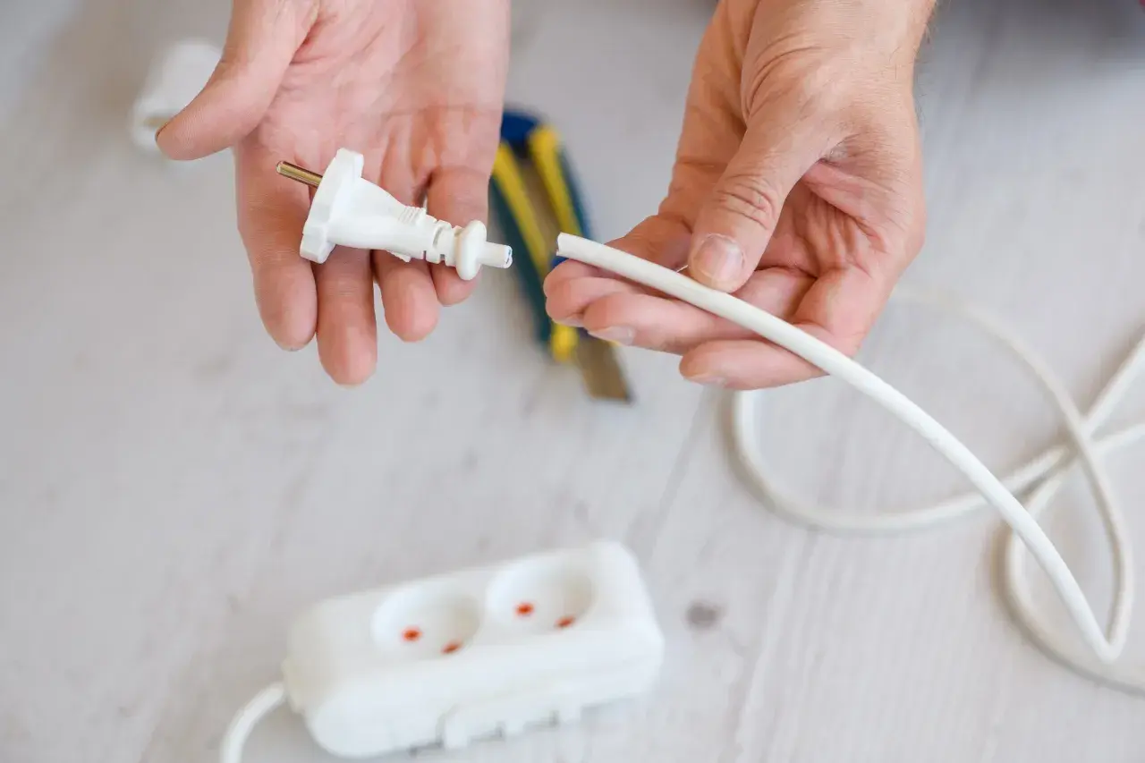 Dłonie przygotowują kabel do podłączenia do wtyczki z uziemieniem. W tle widać listwę zasilającą.