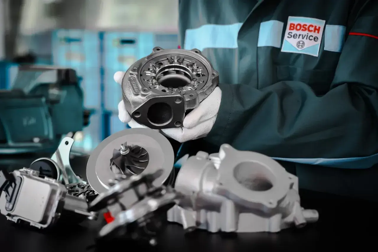 Mechanik w rękawiczkach prezentuje części turbosprężarki, w tym wirnik i obudowę. Na jego kombinezonie widnieje logo Bosch Service. P0299.